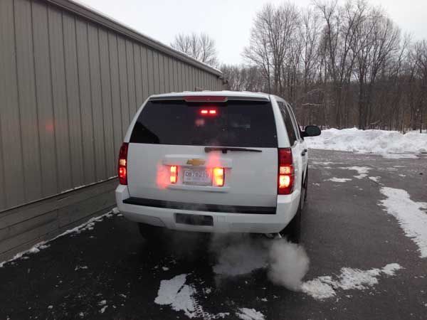 White SUV with tail lights on, exhaling smoke on a snowy driveway.