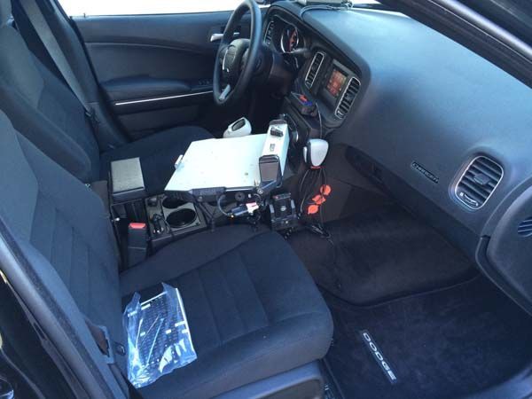 Interior of a police car; driver's side. Black dashboard, center console with equipment, black seats, and steering wheel.