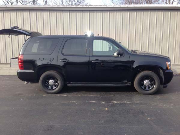 Black police SUV with emergency lights, parked in front of a gray building. The back hatch is open.
