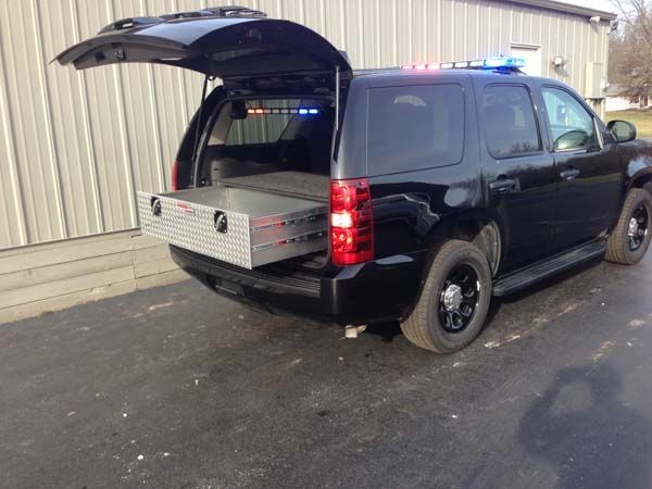 Black SUV with open tailgate, a metal storage box slides out; emergency lights on.