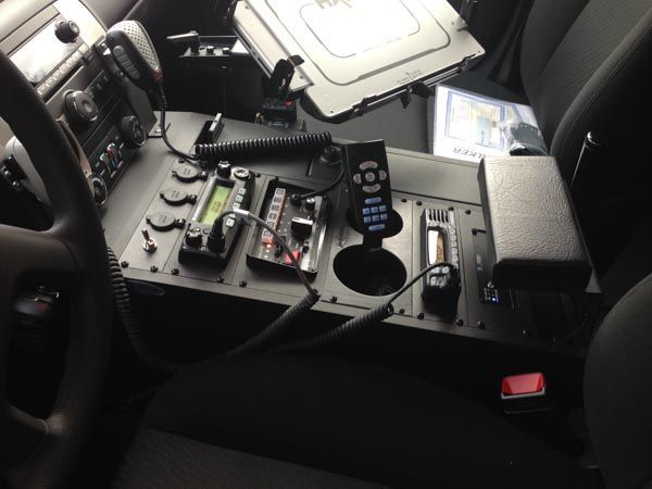 Interior of a police vehicle with a center console containing radio equipment, a cup holder, and a small control screen.