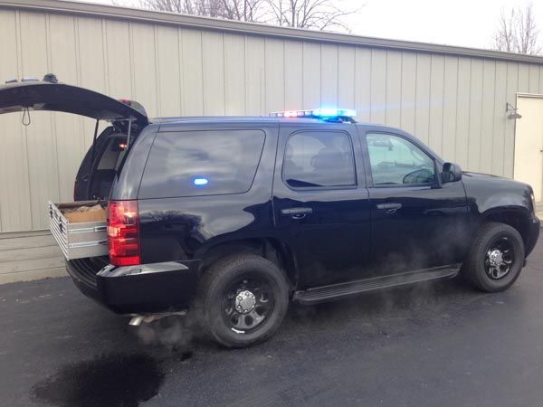 Black SUV with flashing blue/red lights, rear hatch open, smoke from exhaust, parked outside a building.