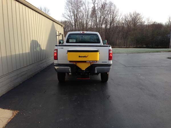 A silver pickup truck with a yellow salt spreader is parked on black pavement next to a building.