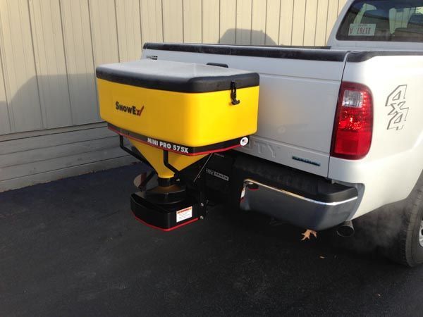 Yellow and black snow spreader attached to the back of a white pickup truck.