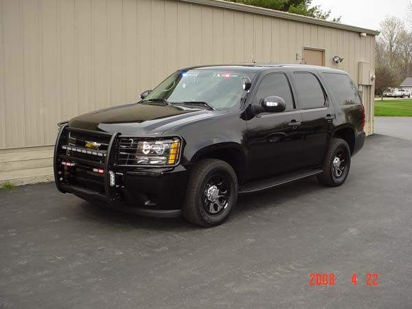 Black Chevrolet Tahoe SUV with push bar, emergency lights, parked outside a tan building.