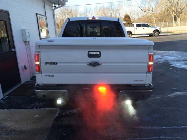 White Ford F-150 truck with exhaust emitting red and white smoke. Tail lights illuminated. Parked outside.