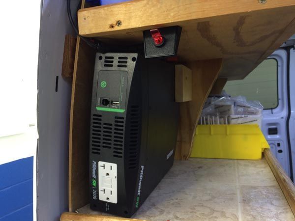 Black inverter with power outlets mounted under a wooden shelf in a van.