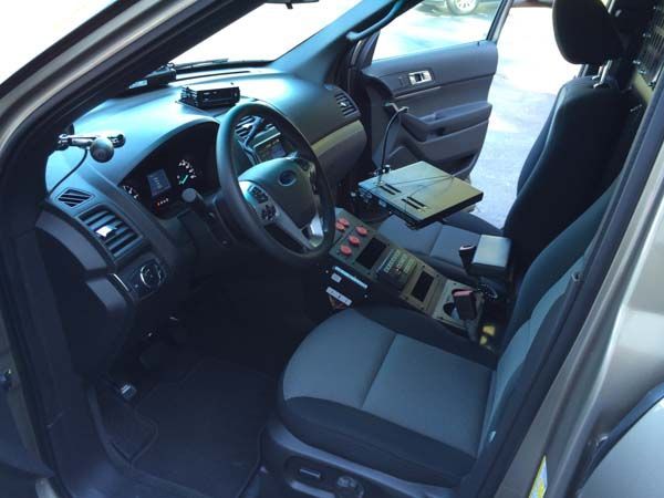 Interior view of a police car with control consoles, computer, and seats. The door is open.
