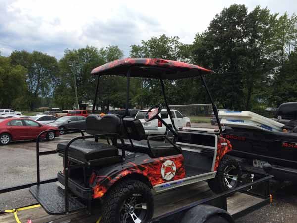Custom golf cart with flame design and black wheels parked on a trailer in a parking lot.