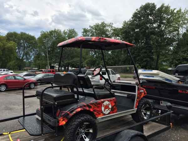 Custom golf cart with flame design on a trailer, parked outside on a cloudy day.