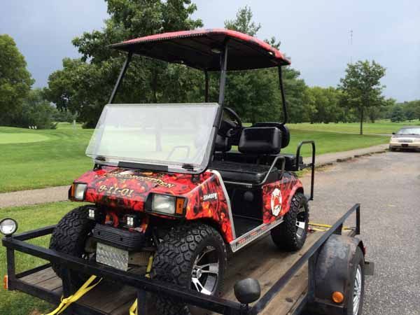 Red golf cart on a trailer, with a camouflage design, parked near a golf course.