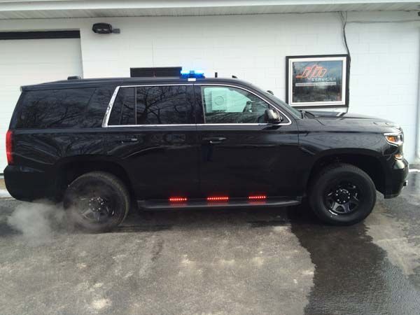 Black SUV with emergency lights, parked in front of a building, exhaust visible.