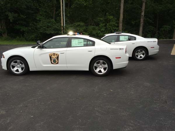 Two white police cars parked on asphalt. A wooded area is in the background.