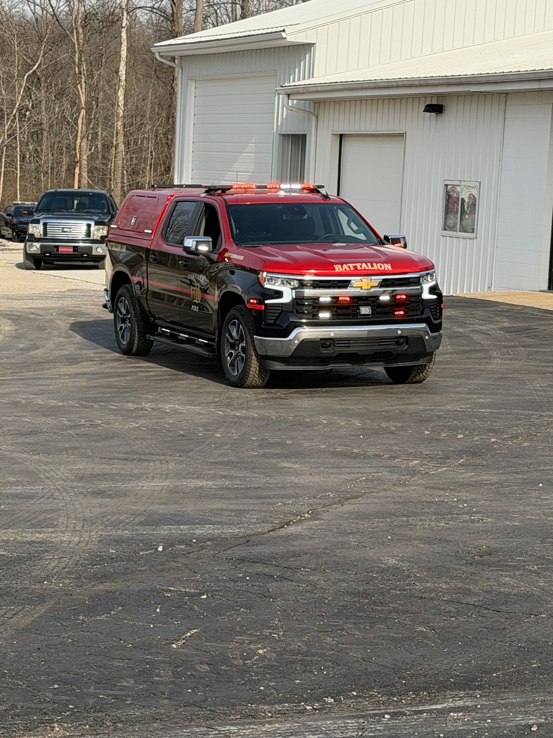 Red SUV with roof lights parked on a gravel lot beside a building, with another vehicle behind it.