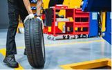 Mechanic holding a tire in a garage, near a red tool cart and a car lift.