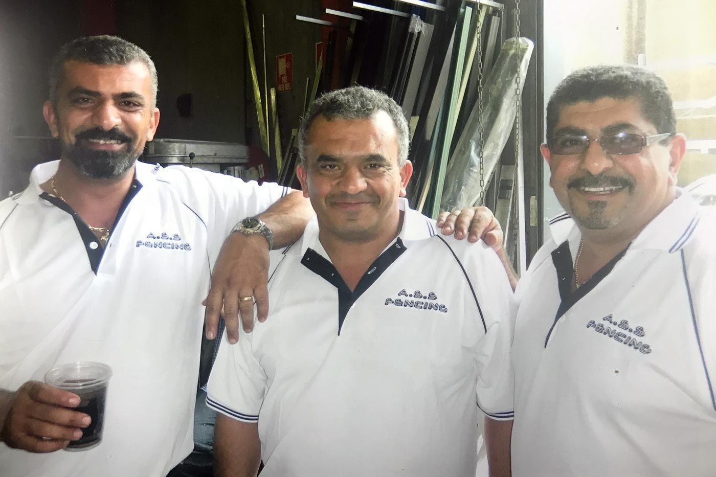 Three men in matching white shirts pose, smiling. The man on the left holds a drink.
