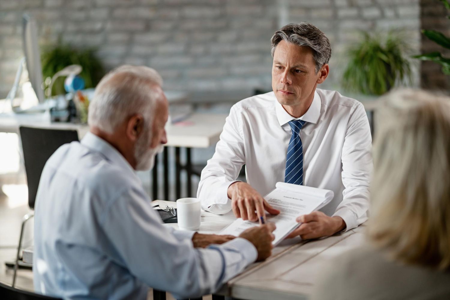 Three people in a meeting, with one man showing documents across a table in an office.