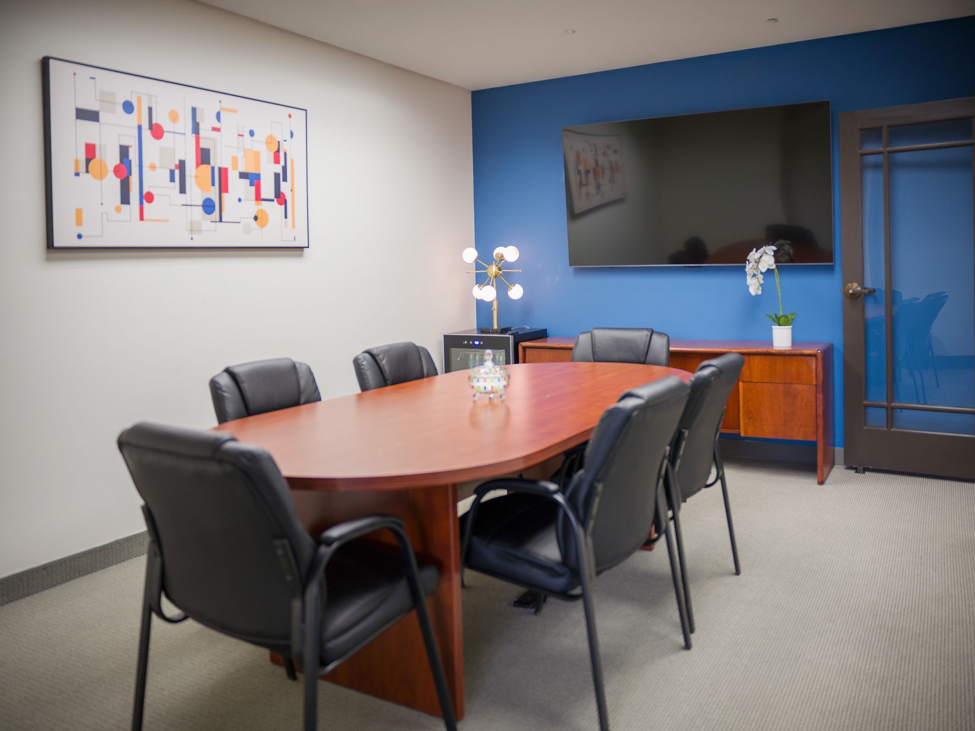 Conference room with oval table, black chairs, wall-mounted TV, and blue accent wall.