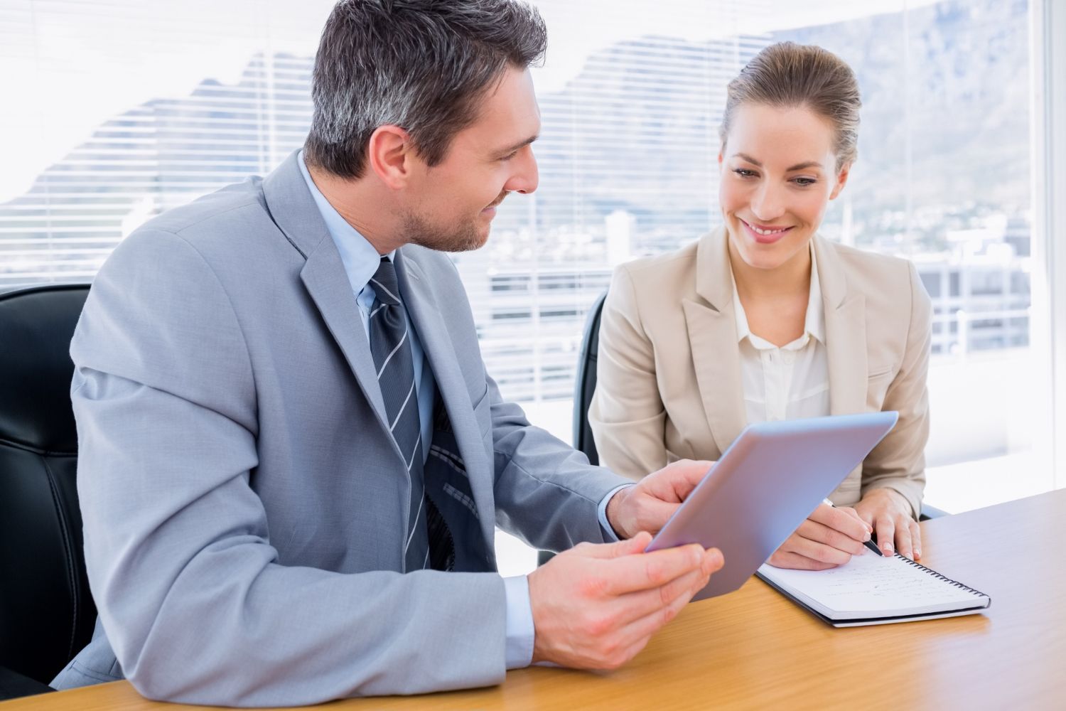 Two coworkers reviewing a tablet in a bright office, with a notebook on the desk.