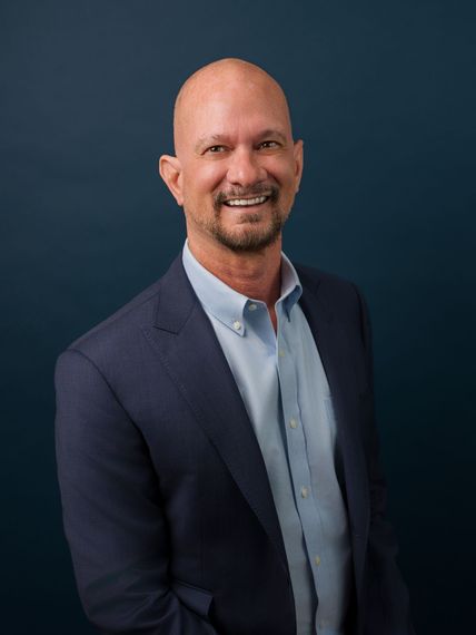 Smiling man in a navy blazer and light blue shirt against a dark blue studio background