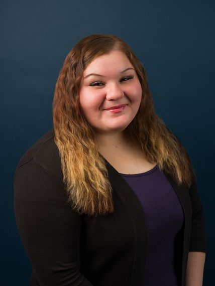 Studio portrait of a smiling person with curly hair, wearing a black cardigan over a purple top, against a blue backdrop