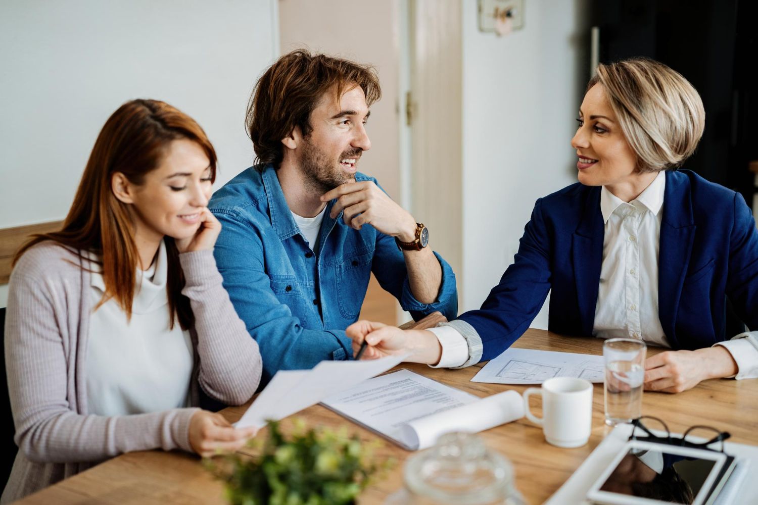 Three people in a meeting smiling and reviewing papers at a table with a laptop and coffee mugs
