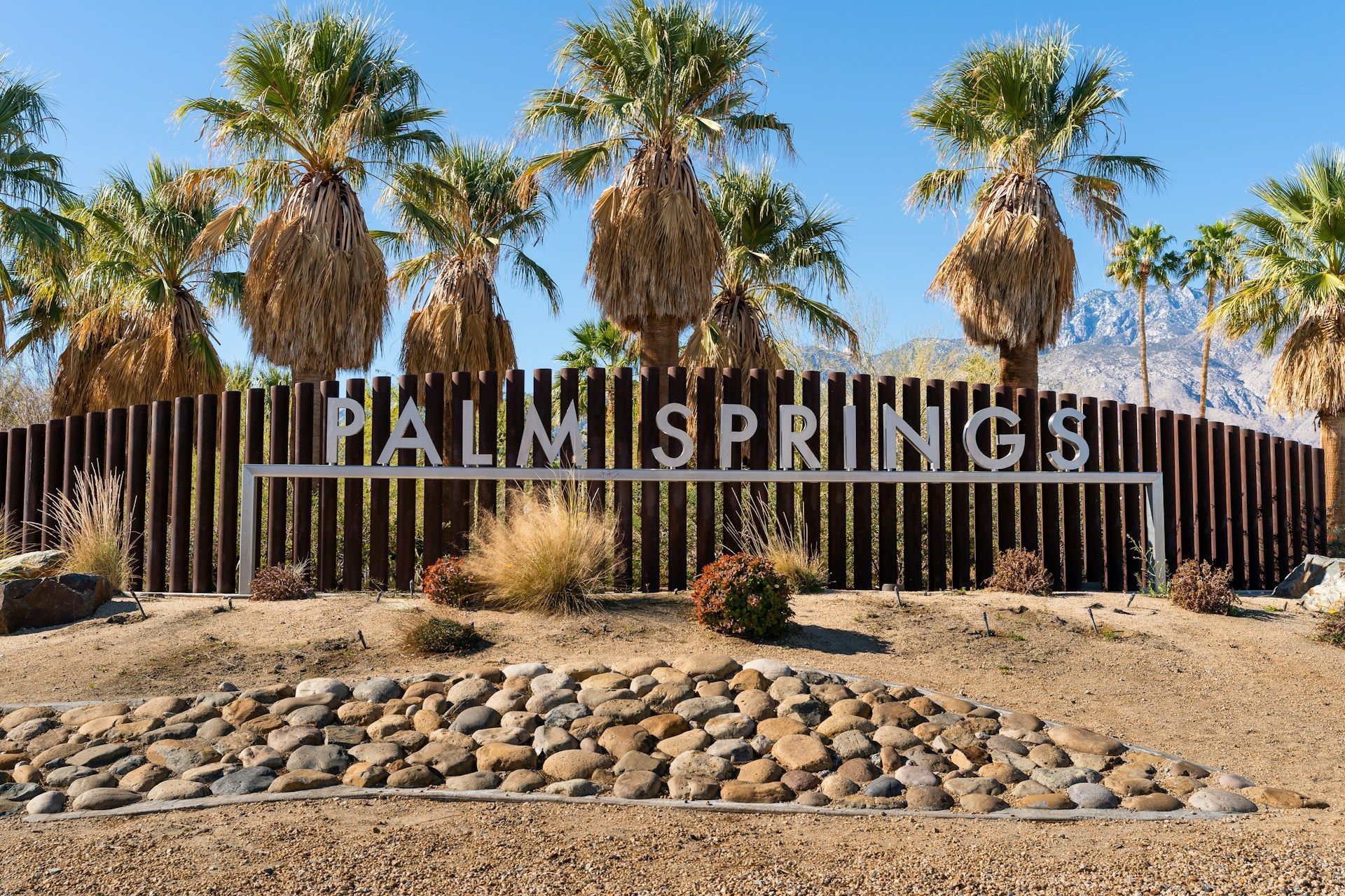 Palm Springs entrance sign with palm trees, desert landscaping, and rocks under a clear blue sky