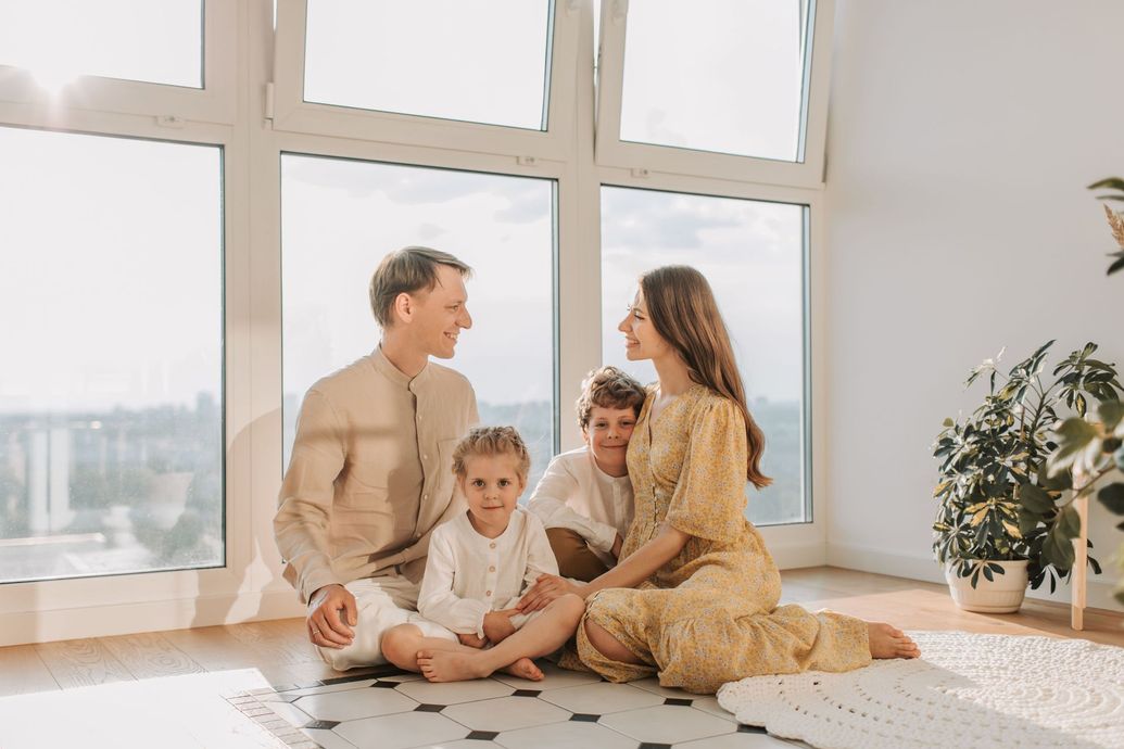 Family sitting on a rug by large windows in a bright living room, sharing a warm moment