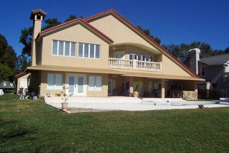 Beige stucco house with red trim, balcony, chimney, and large patio on a sunny day.