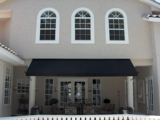 Beige house with arched windows above a black awning covering an outdoor patio with seating.