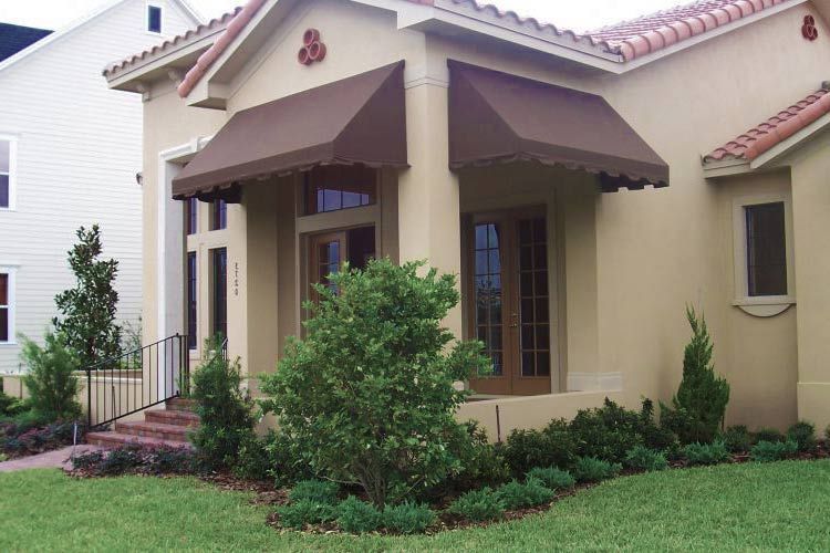 Beige house with brown awnings, windows, and landscaping, and a green lawn.