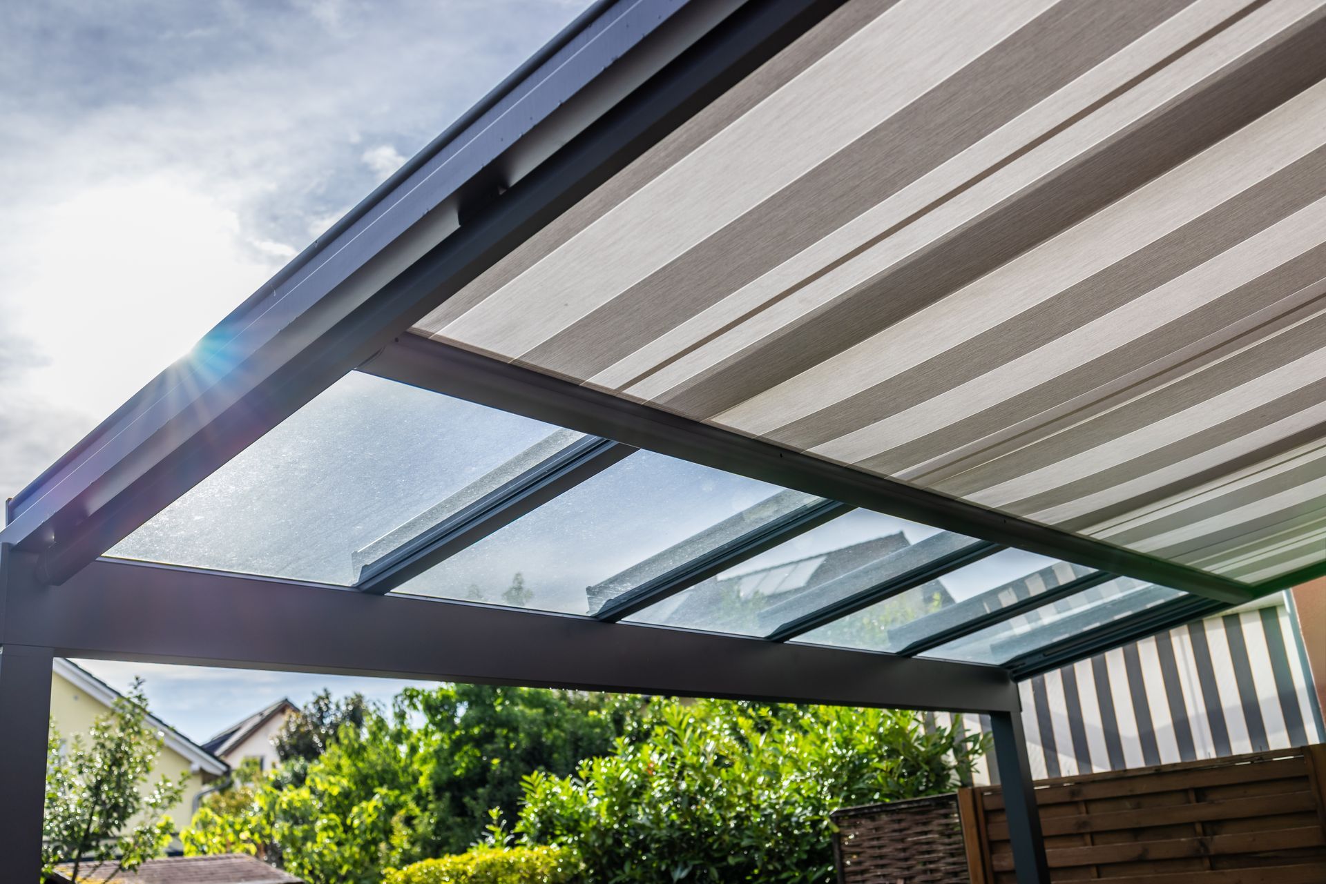 Glass-roof pergola with striped awning panels over a backyard patio in sunlight.