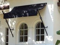 Black metal window awning mounted above arched windows on a white stucco exterior. Black metal window awning mounted above arched windows on a white stucco exterior.