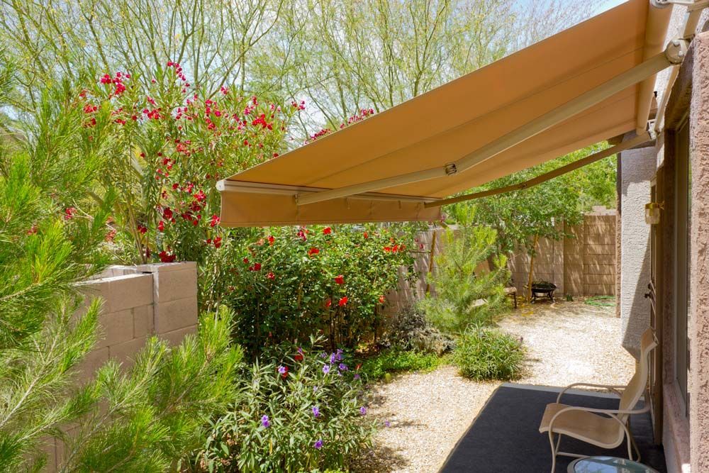 Beige awning extends over a patio area with a stone path, plants, and a tan wall.