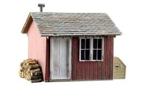 Small red wooden shed with a gray shingled roof, white door, and stacked firewood beside it.