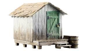 Weathered white wooden shack with a green door and tin roof, raised on stilts beside barrels.