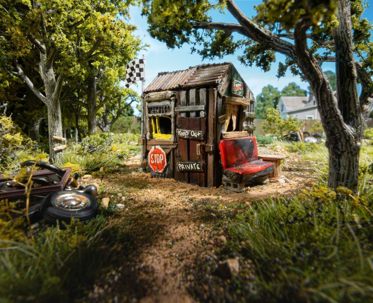 Small rustic roadside hut with a red motorbike under trees, surrounded by grass and dirt path.