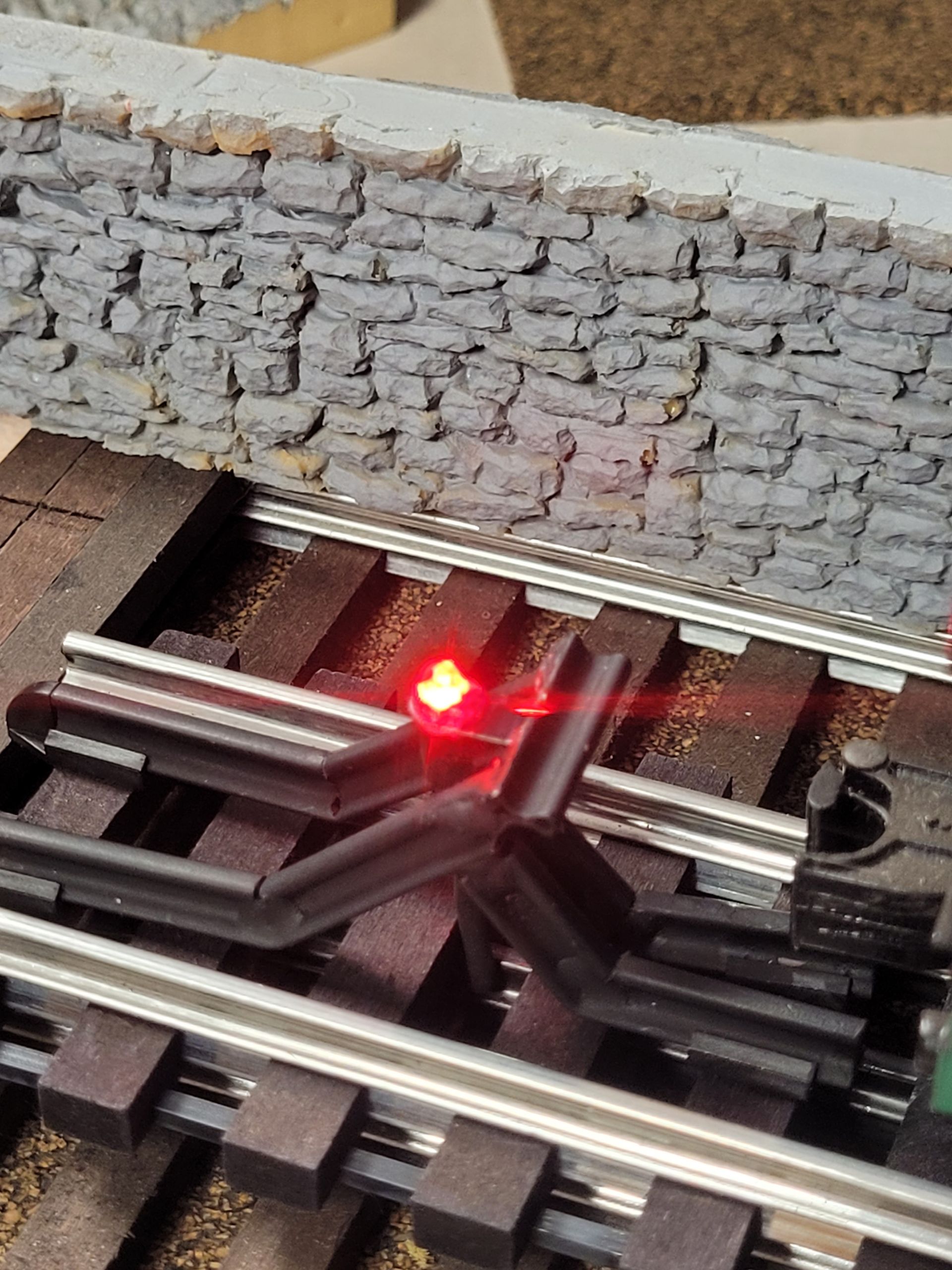 Toy railroad track layout with a bright red signal light beside a gray stone wall