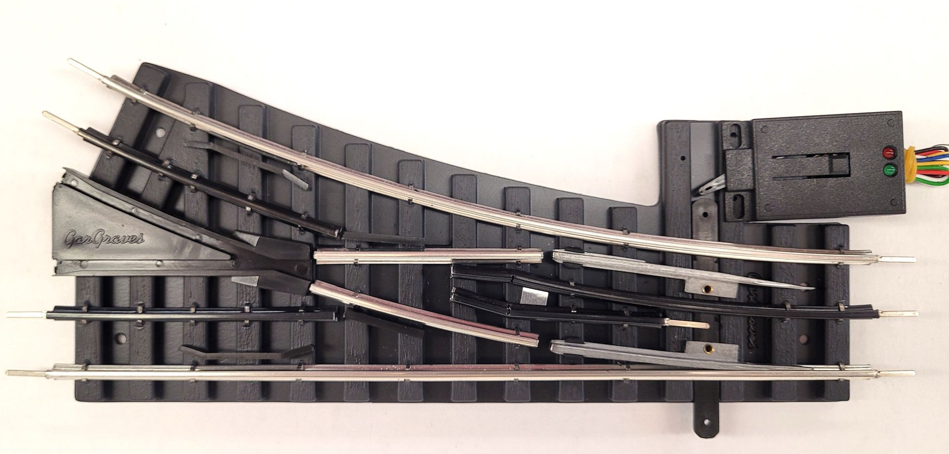 Model railway switch track with a dark control box and colored wires on a white background