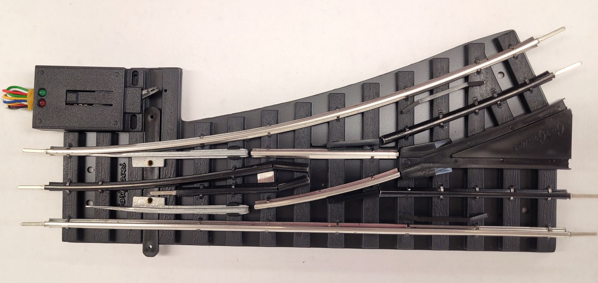 Black model train turnout switch track with a motorized mechanism on the left.