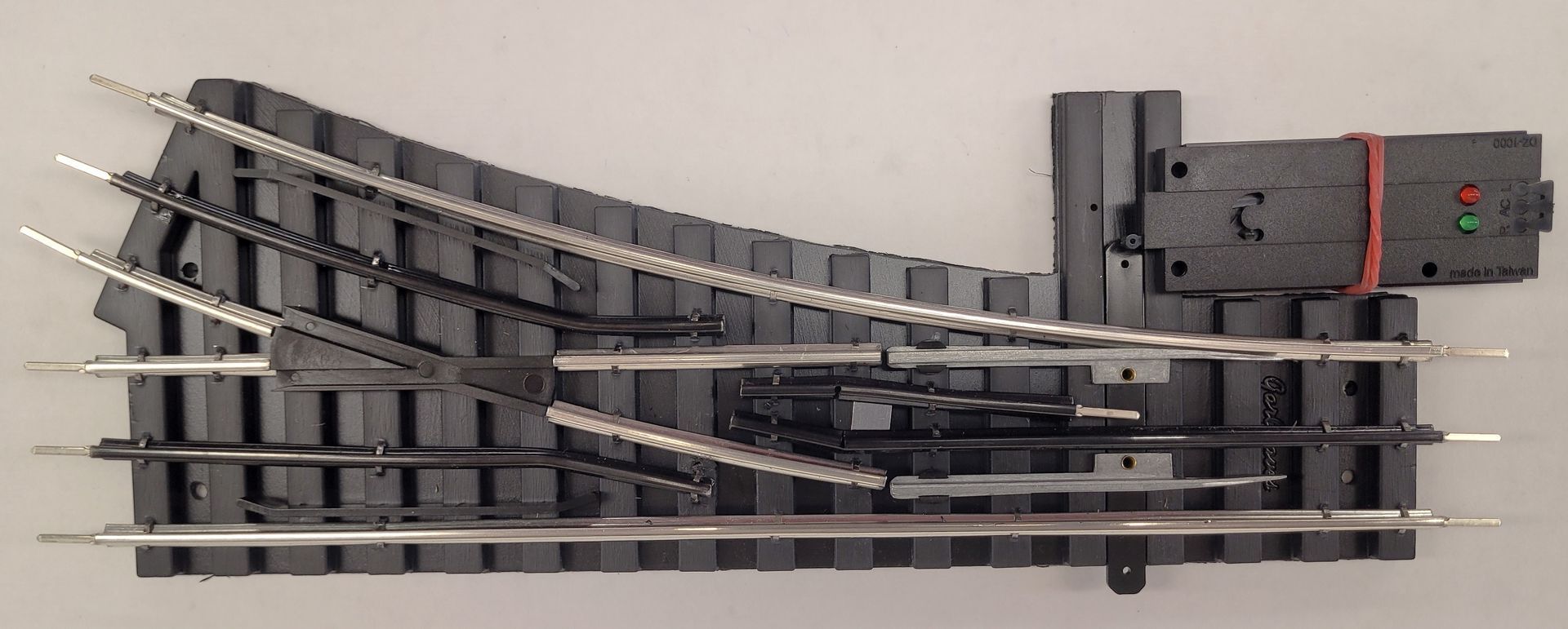 Model railway turnout switch with black tracks and a gray motor housing on a white background