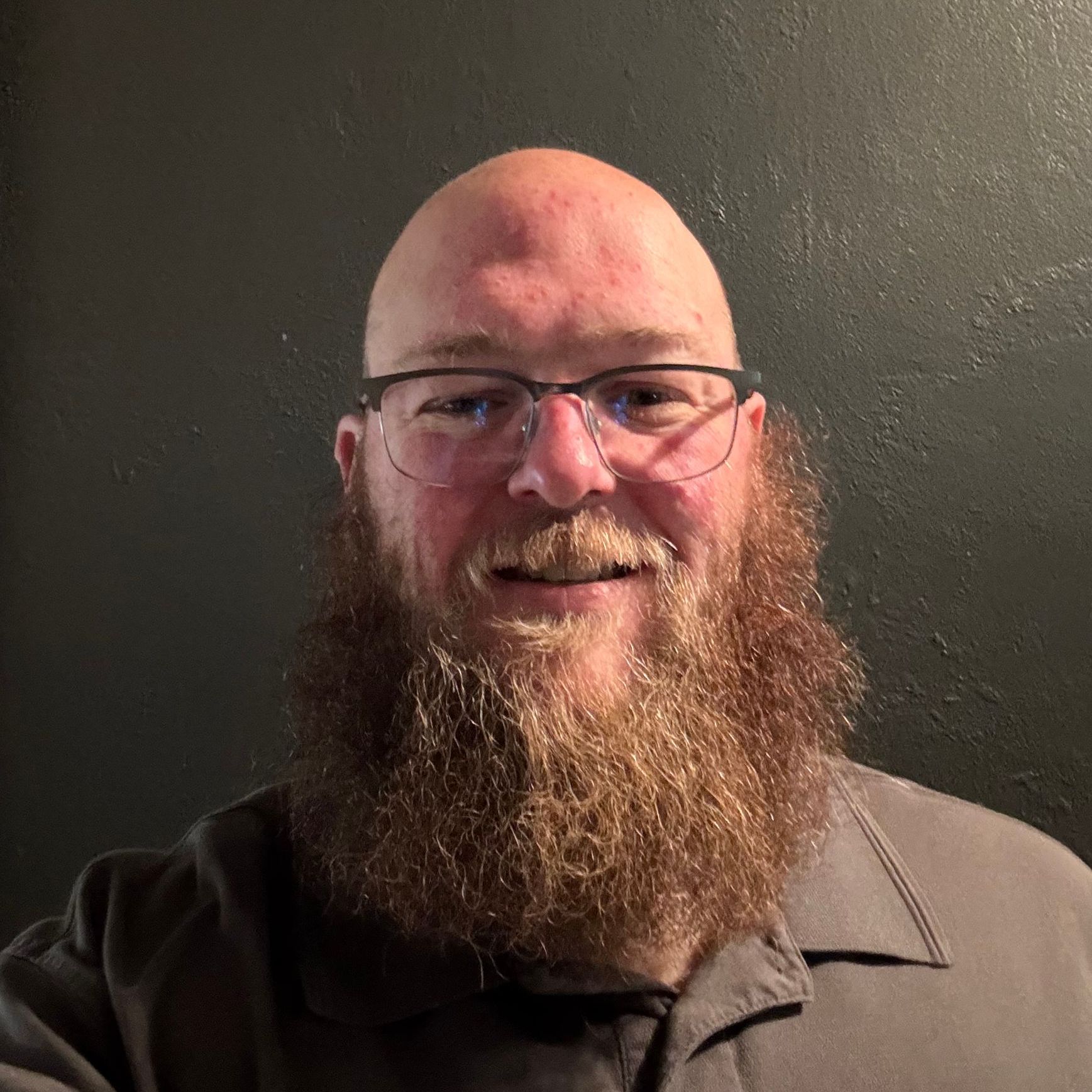 Bald man with glasses and long beard smiles, grey wall background.