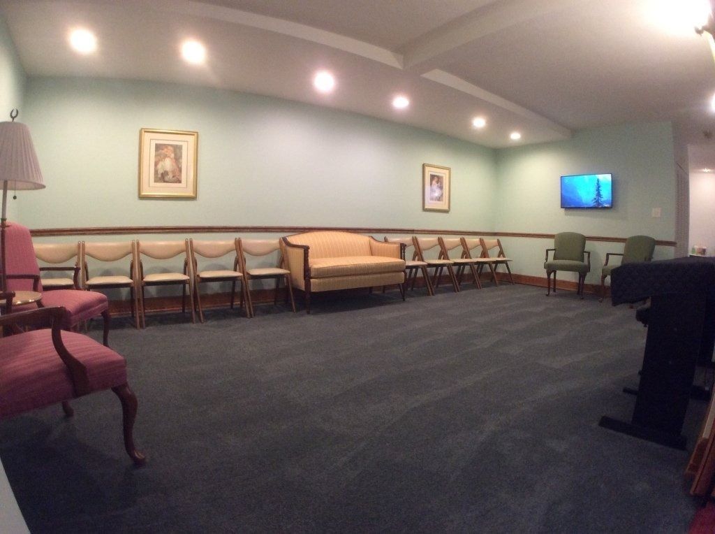 Quiet waiting room with chairs, beige couch, wall art, TV, and dark carpet under recessed lights.