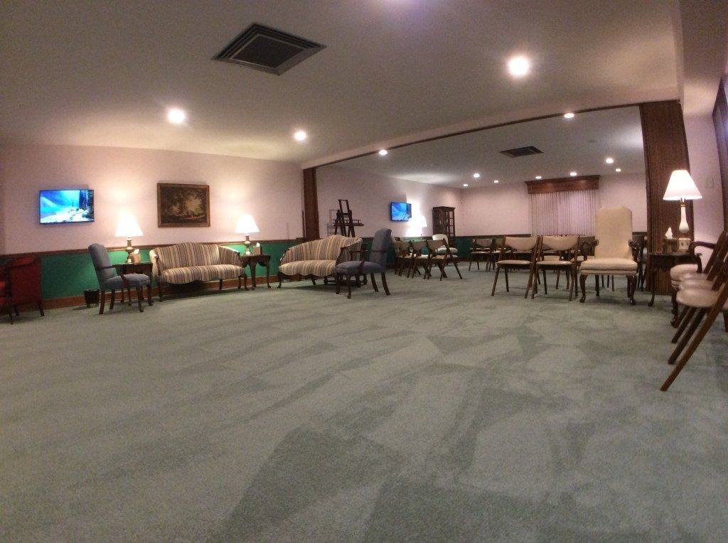 Empty carpeted lounge with tables, chairs, and warm ceiling lights; TV mounted on the left wall.