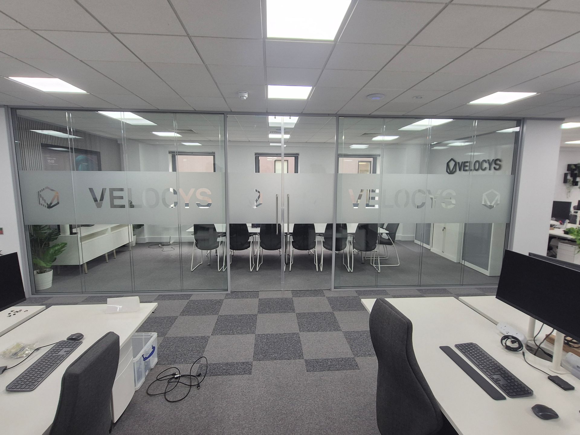 Empty modern office with glass walls, desks, computers, and gray carpet tiles.