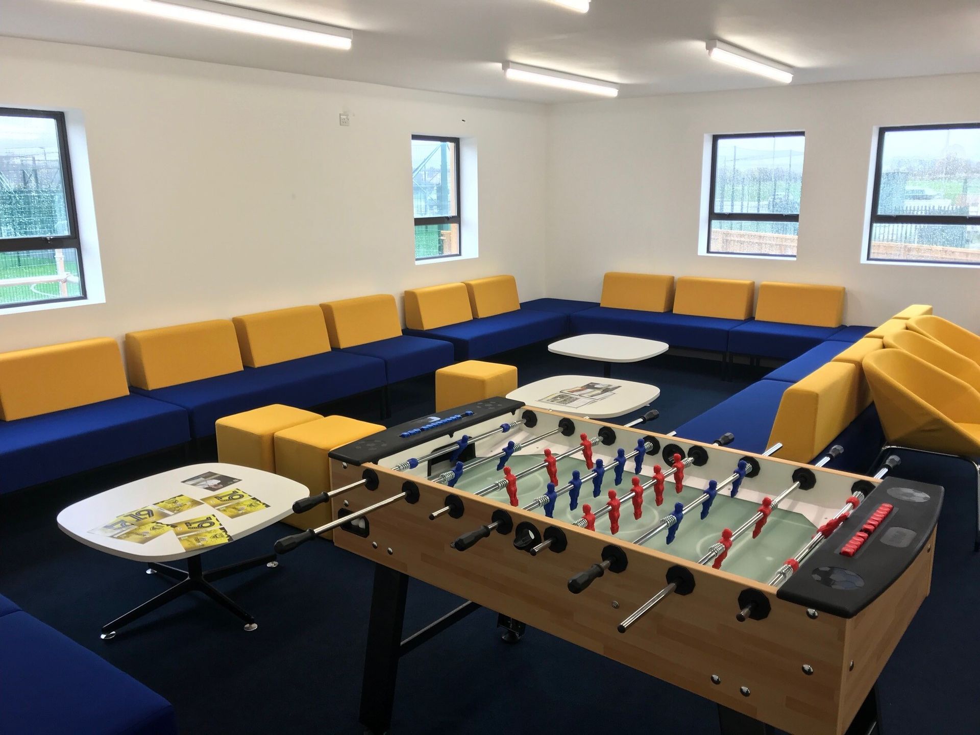 Recreation room with foosball table, round tables, blue sofas, and yellow cushions under bright windows