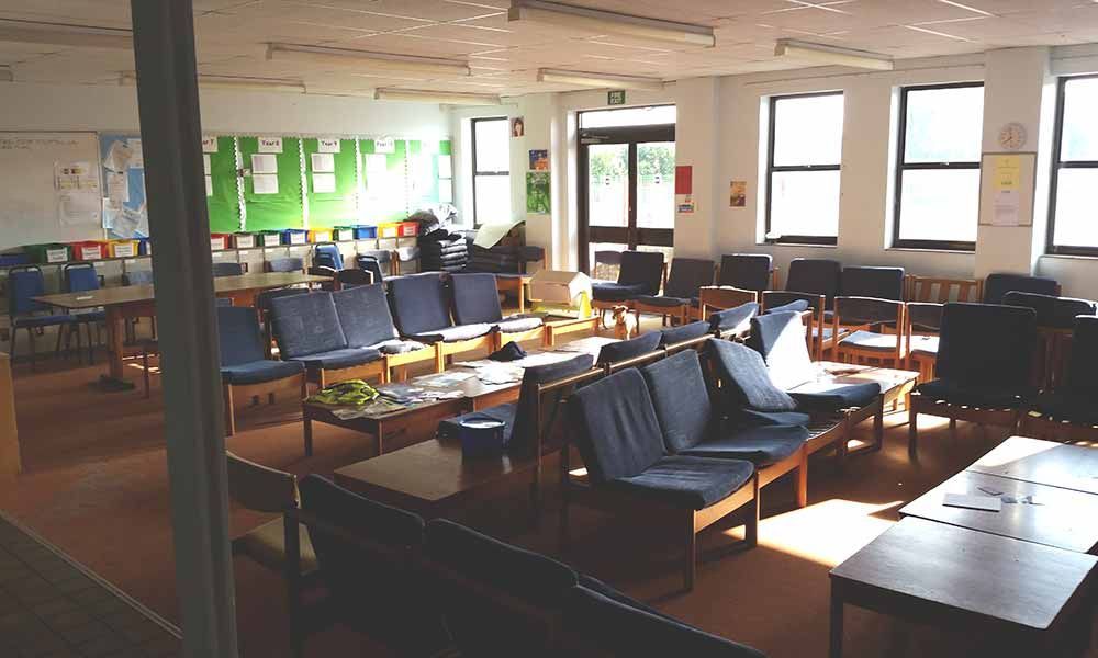 Empty classroom with rows of desks, blue chairs, and green bulletin boards near large windows.
