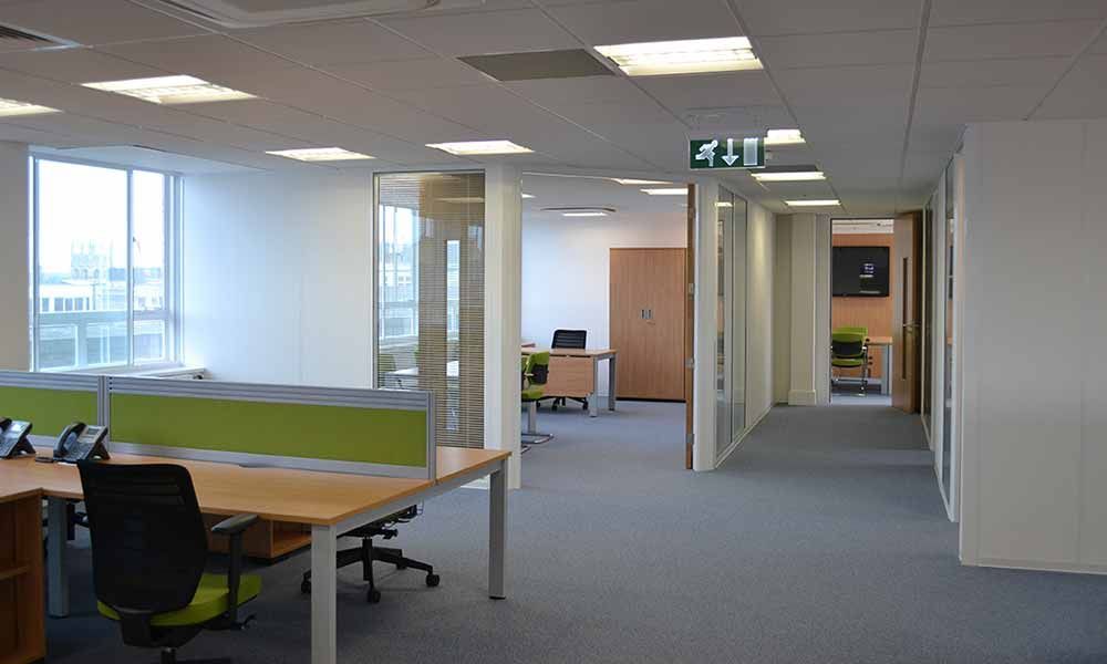 Empty office with desks, chairs, carpeted floor, and large windows in bright daylight.