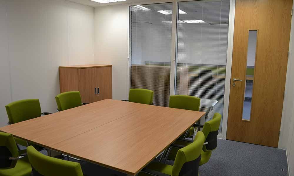 Small conference room with a wood table, green chairs, glass partition, and wooden door