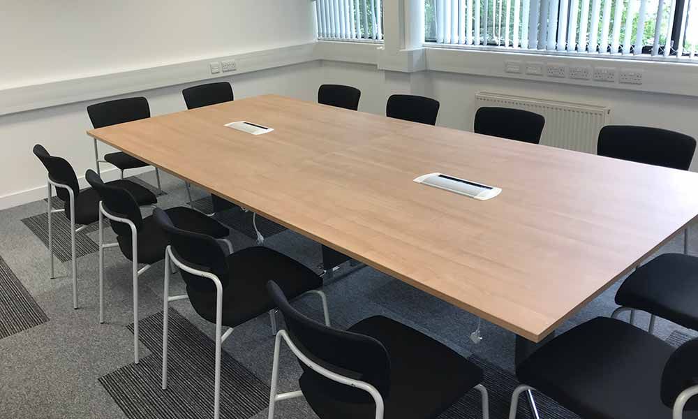 Empty conference room with a large wooden table and black chairs under bright windows.